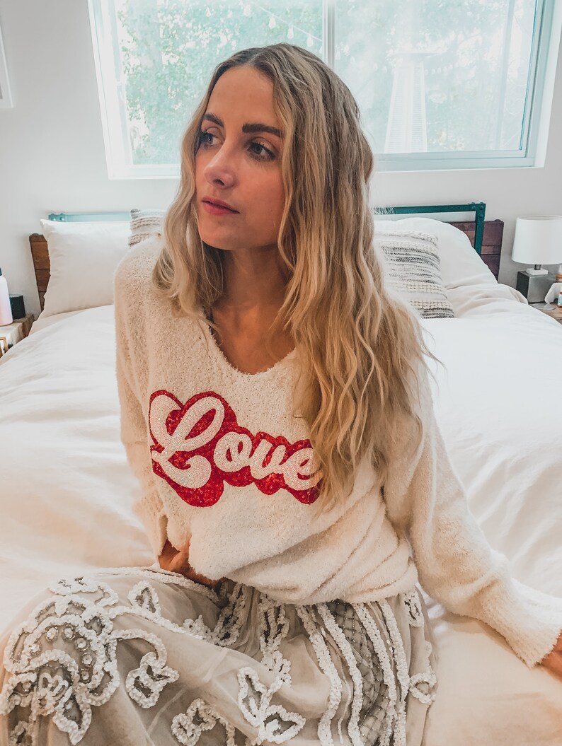 May include: A white fuzzy sweater with a red "Love" graphic. The sweater is worn by a person sitting on a bed with a white bedspread. The person is wearing a white skirt with a lace design.