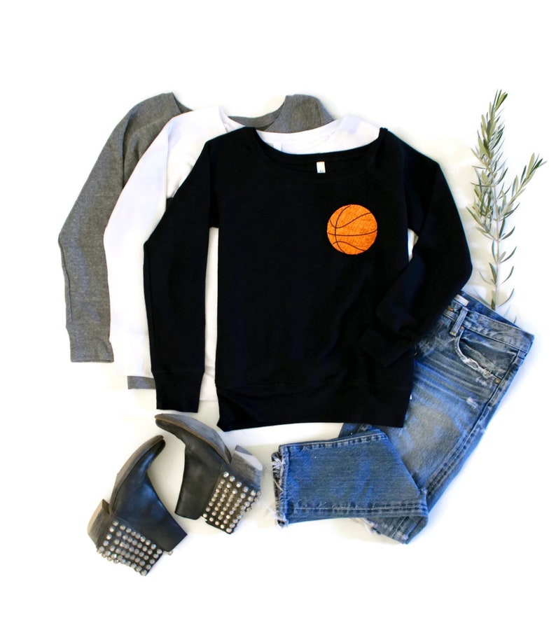 May include: A black long-sleeved sweatshirt with a basketball graphic on the front. The sweatshirt is layered over a white long-sleeved shirt and a grey long-sleeved shirt. The sweatshirt is paired with blue jeans and black studded ankle boots.
