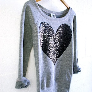 May include: A gray sweatshirt with a large black sequined heart on the front. The sweatshirt has long sleeves and a relaxed fit.