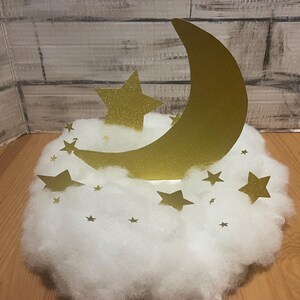 Pode incluir: Uma lua crescente dourada com glitter e estrelas em uma nuvem branca e fofa. A lua e as estrelas s&atilde;o cortadas em papel e t&ecirc;m um acabamento brilhante.