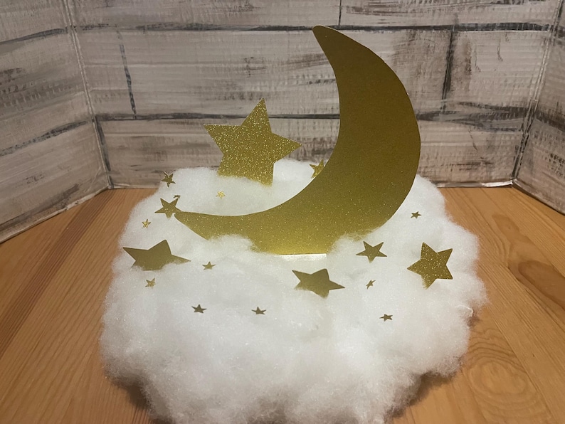 Pode incluir: Uma lua crescente dourada com glitter e estrelas em uma nuvem branca e fofa. A lua e as estrelas s&atilde;o cortadas em papel e t&ecirc;m um acabamento brilhante.