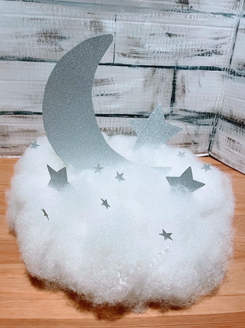 Pode incluir: Uma lua prateada com glitter e estrelas em uma nuvem branca e fofa. A lua tem a forma de uma lua crescente e as estrelas t&ecirc;m cinco pontas.