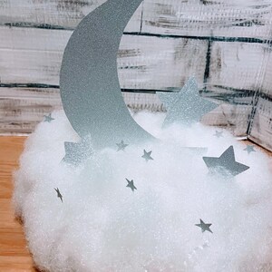 Pode incluir: Uma lua prateada com glitter e estrelas em uma nuvem branca e fofa. A lua tem a forma de uma lua crescente e as estrelas t&ecirc;m cinco pontas.