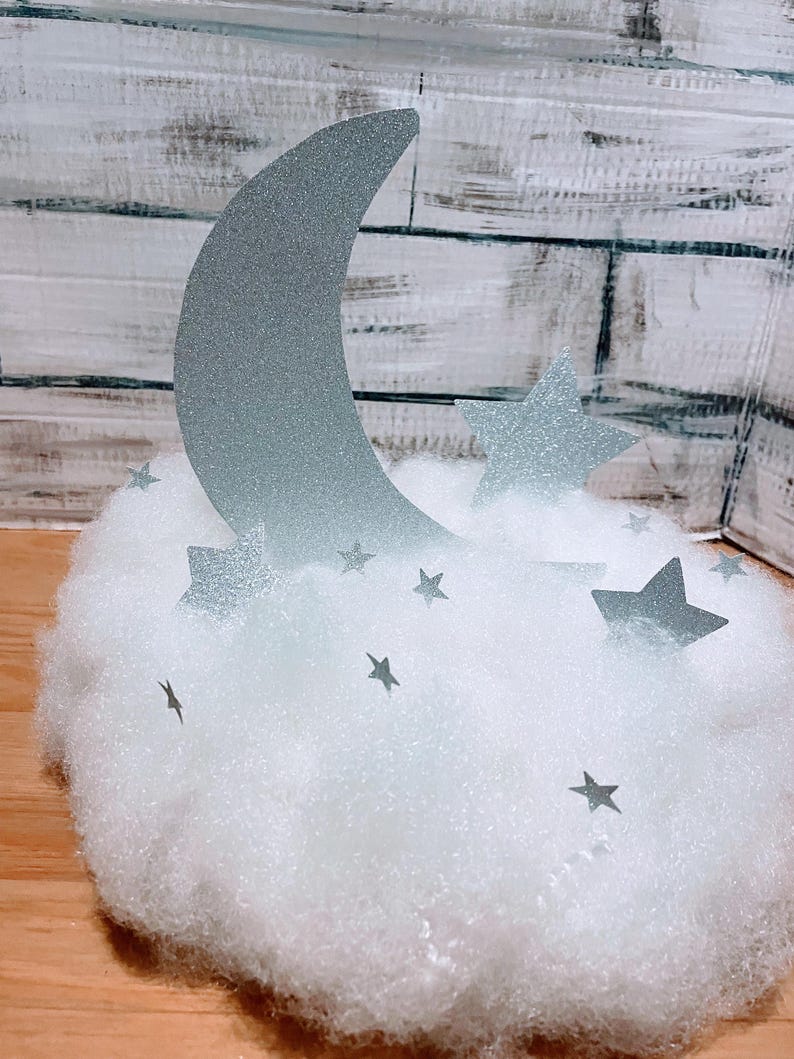Pode incluir: Uma lua prateada com glitter e estrelas em uma nuvem branca e fofa. A lua tem a forma de uma lua crescente e as estrelas t&ecirc;m cinco pontas.