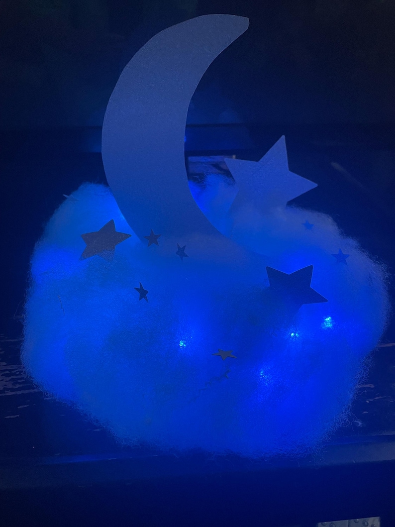 Pode incluir: Uma luz noturna em forma de nuvem azul com uma lua crescente branca e estrelas. A luz &eacute; iluminada com luzes LED azuis.