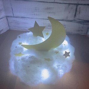 Pode incluir: Uma nuvem branca e fofa com uma lua crescente amarela e uma estrela amarela no topo. A nuvem &eacute; iluminada por luzes brancas.