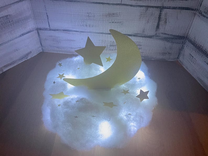 Pode incluir: Uma nuvem branca e fofa com uma lua crescente amarela e uma estrela amarela no topo. A nuvem &eacute; iluminada por luzes brancas.