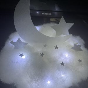 Pode incluir: Uma nuvem branca e fofa com uma lua crescente branca e estrelas brancas, iluminadas por pequenas luzes LED brancas.
