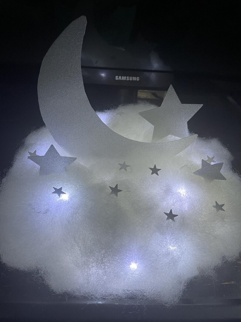 Pode incluir: Uma nuvem branca e fofa com uma lua crescente branca e estrelas brancas, iluminadas por pequenas luzes LED brancas.