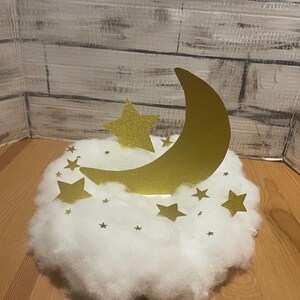 Pode incluir: Uma decora&ccedil;&atilde;o de lua crescente dourada e estrela em uma nuvem branca e fofa. A lua e as estrelas s&atilde;o feitas de papel com glitter.