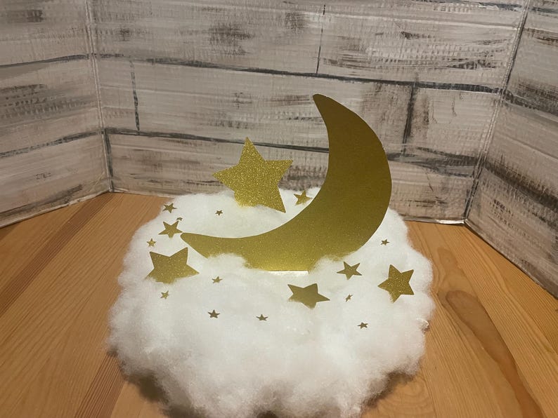 Pode incluir: Uma decora&ccedil;&atilde;o de lua crescente dourada e estrela em uma nuvem branca e fofa. A lua e as estrelas s&atilde;o feitas de papel com glitter.