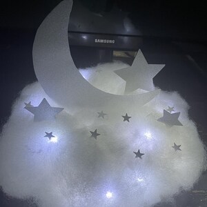Pode incluir: Uma luz branca em forma de nuvem com recortes em forma de lua crescente e estrelas. A luz &eacute; iluminada por luzes LED brancas.