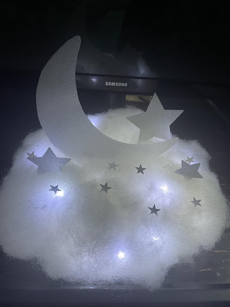 Pode incluir: Uma luz branca em forma de nuvem com recortes em forma de lua crescente e estrelas. A luz &eacute; iluminada por luzes LED brancas.