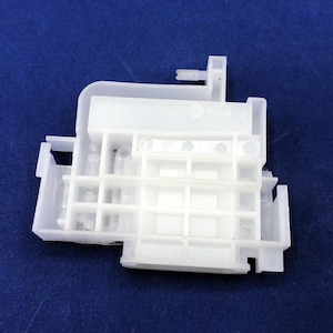 May include: A white plastic component with a complex, geometric design. The piece features a series of rectangular and square openings, along with protruding tabs and a grid-like structure. The object is set against a solid blue background.