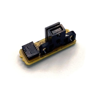 Puede incluir: Primer plano de una pequeña placa de circuito electrónico con un conector negro y un sensor negro. La placa es amarilla y tiene marcas negras.