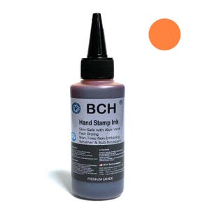 Puede incluir: Una botella de tinta para sellos BCH con tapa y boquilla negras. La etiqueta indica que es segura para la piel con aloe vera, de secado rápido y no tóxica. Un círculo naranja está en la esquina superior derecha.