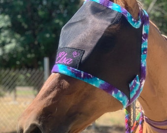 May include: A brown horse wearing a black fly mask with a purple and blue tie-dye trim. The mask has a black patch with the name "Elsa" embroidered in pink.