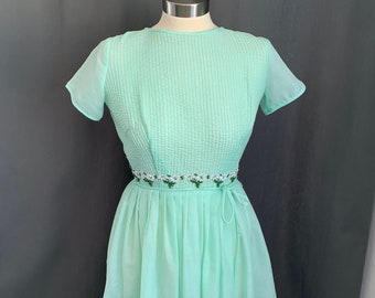 May include: A mint green dress with short sleeves and a white floral embroidered belt. The dress has a gathered skirt and a fitted bodice.