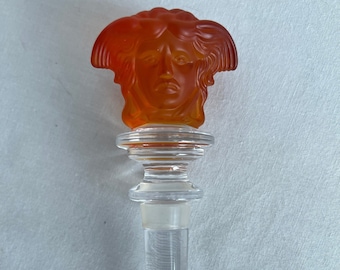 May include: Orange glass bottle stopper with a detailed head of Medusa design. The stopper is clear glass with a textured base.