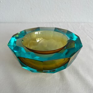 Op de afbeelding: Een gefacetteerde glazen kom met een tealblauwe buitenkant en een gele binnenkant. De kom heeft een geometrische vorm en is perfect om kleine voorwerpen te presenteren.