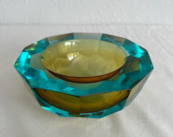 May include: A faceted glass bowl with a teal blue exterior and a yellow interior. The bowl has a geometric shape and is perfect for displaying small items.