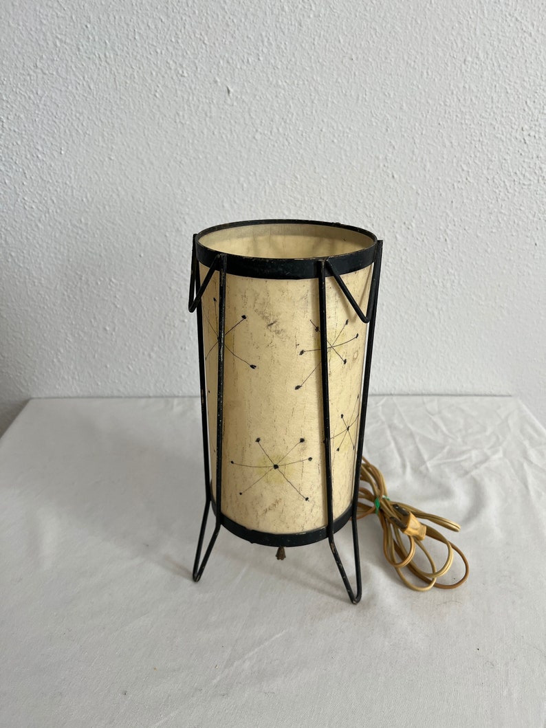 May include: A black metal table lamp with a white paper shade featuring a geometric pattern of black lines and dots. The lamp has a cord and plug.