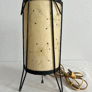 May include: A mid-century modern table lamp with a black metal frame and a white shade with a geometric pattern. The lamp has a cord and plug.