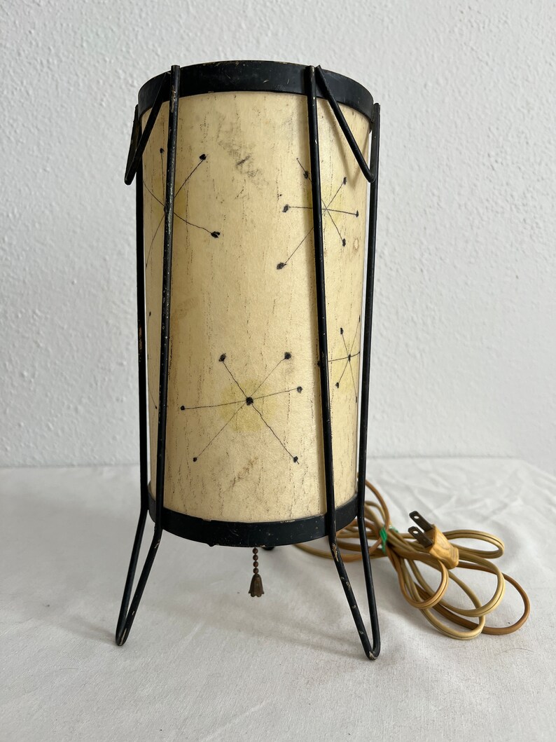 May include: A mid-century modern table lamp with a black metal frame and a white shade with a geometric pattern. The lamp has a cord and plug.