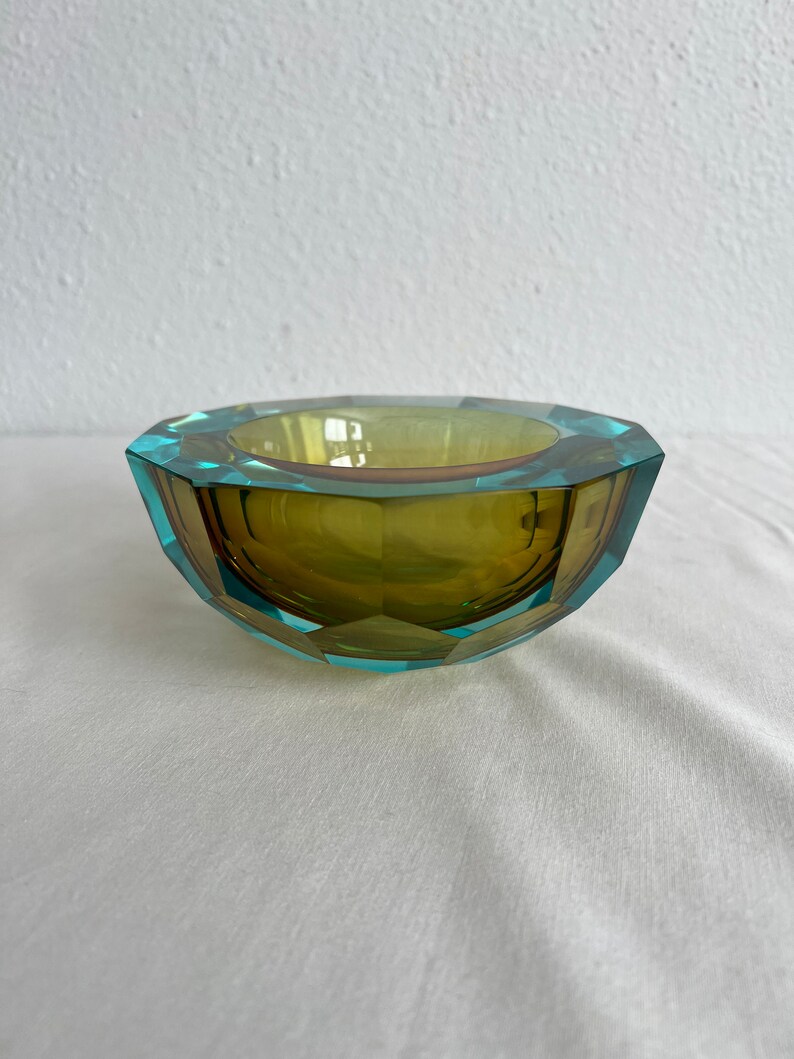 Op de afbeelding: Een gefacetteerde glazen kom met een groen en blauw kleurenschema. De kom heeft een geometrische vorm en is gemaakt van helder glas.