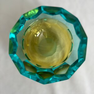 Op de afbeelding: Een gefacetteerde glazen kom met een tealblauwe en groene buitenkant en een gele binnenkant.