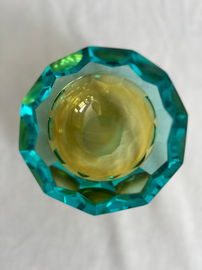 Op de afbeelding: Een gefacetteerde glazen kom met een tealblauwe en groene buitenkant en een gele binnenkant.