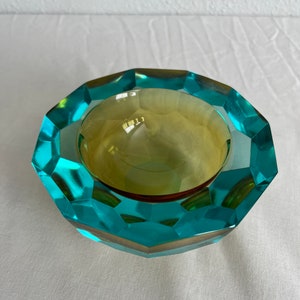 Op de afbeelding: Een asbak van turquoise en amberkleurig glas met een facetgeslepen ontwerp. De asbak is rond en heeft een ondiepe kom.