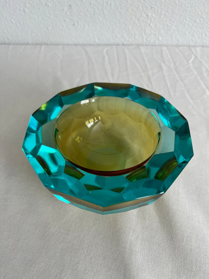 Op de afbeelding: Een asbak van turquoise en amberkleurig glas met een facetgeslepen ontwerp. De asbak is rond en heeft een ondiepe kom.