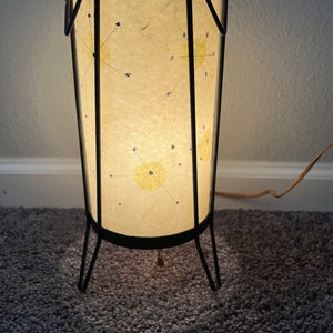 May include: A mid-century modern table lamp with a black metal frame and a white paper shade with a starburst design. The lamp is on and casting a warm glow.