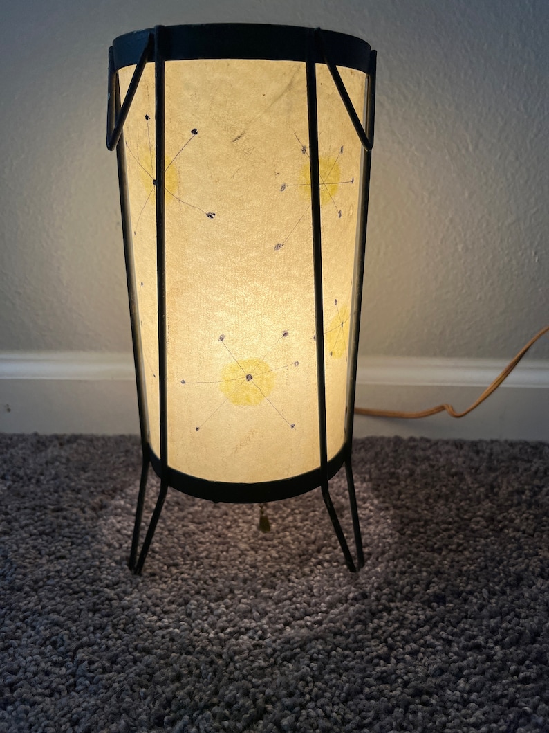 May include: A mid-century modern table lamp with a black metal frame and a white paper shade with a starburst design. The lamp is on and casting a warm glow.