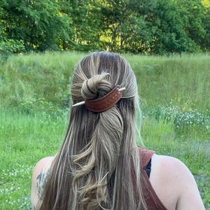 May include: A brown leather hair stick with a metal pin. The stick is being used to hold a woman's hair in a bun.