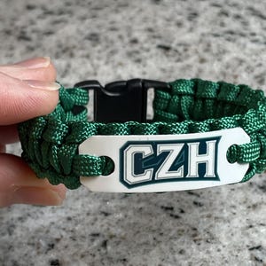 Peut inclure: Un bracelet en paracorde vert doté d'une plaque métallique rectangulaire blanche avec les lettres CZH imprimées en bleu foncé avec un contour blanc. Le bracelet comprend une boucle de fermeture latérale en plastique noir.