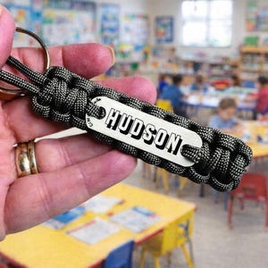 May include: A black and grey paracord keychain featuring a rectangular silver-toned metal tag engraved with the name HUDSON in bold black capital letters. The braided cord is attached to a circular metal split ring for keys.