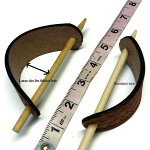 May include: Two leather hair sticks, one standard size and one larger size for thicker hair. The sticks are made of wood and the leather is brown. A measuring tape is used to show the size difference. "Made in China" and "60 in" are printed on the tape.