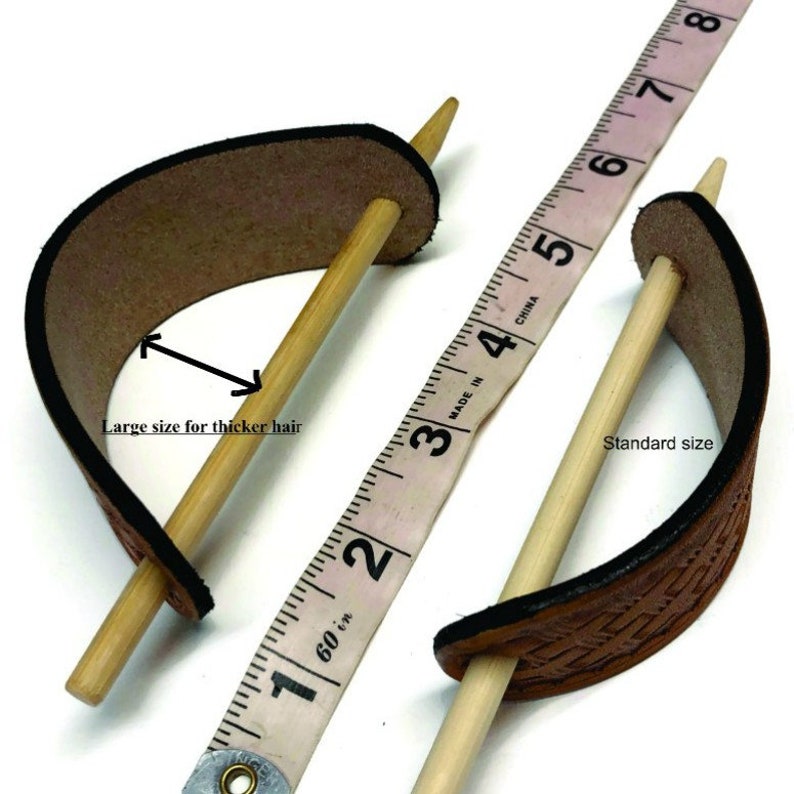 May include: Two leather hair sticks, one standard size and one larger size for thicker hair. The sticks are made of wood and the leather is brown. A measuring tape is used to show the size difference. "Made in China" and "60 in" are printed on the tape.