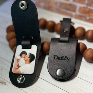 May include: Two black leather keychains with a snap closure. One keychain has a photo insert with a picture of a father and daughter. The other keychain has the word "Daddy" engraved on it.