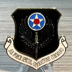 May include: A gold-colored metal coin with a black shield featuring a white star with a red center, a dagger, and wings. The shield is surrounded by a white scroll with the text "Air Force Special Operations Command".