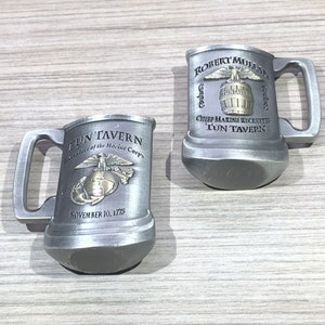 May include: Two silver pewter mugs with a gold eagle and globe design. The mugs feature the text "TUN' TAVERN" and "Birthplace of the Marine Corps" on one mug and "ROBERT MULLAN" and "Chief Marine Recruiter TUN' TAVERN" on the other mug.