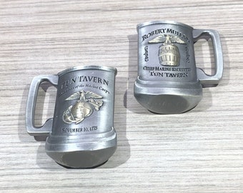 Puede incluir: Dos jarras de peltre plateado con un diseño de águila dorada y globo terráqueo. Las jarras tienen el texto "TUN' TAVERN" y "Birthplace of the Marine Corps" en una jarra y "ROBERT MULLAN" y "Chief Marine Recruiter TUN' TAVERN" en la otra jarra.