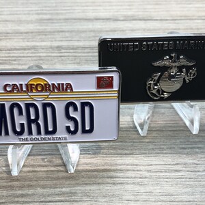 May include: Two metal pins on display stands. The first pin is a black and silver United States Marine Corps pin. The second pin is a white and silver California license plate with the text "MCRD SD" and "The Golden State".