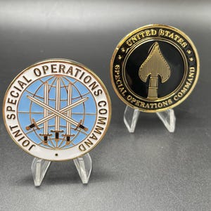 May include: Two gold-colored challenge coins with the Joint Special Operations Command logo. The coin on the left has a blue globe with crossed swords and the text "JOINT SPECIAL OPERATIONS COMMAND". The coin on the right has a black background with a gold spearhead and the text "UNITED STATES SPECIAL OPERATIONS COMMAND".