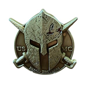 K&ouml;nnte beinhalten: Eine bronzefarbene M&uuml;nze mit einem Spartanerhelm-Design. Die M&uuml;nze tr&auml;gt die Aufschrift "US MC" und "BLOOD BROTHERS". Zwei gekreuzte Schwerter sind ebenfalls vorhanden. Die M&uuml;nze hat ein abgenutztes, gealtertes Aussehen, mit einem Durchmesser von ca. 4 cm.