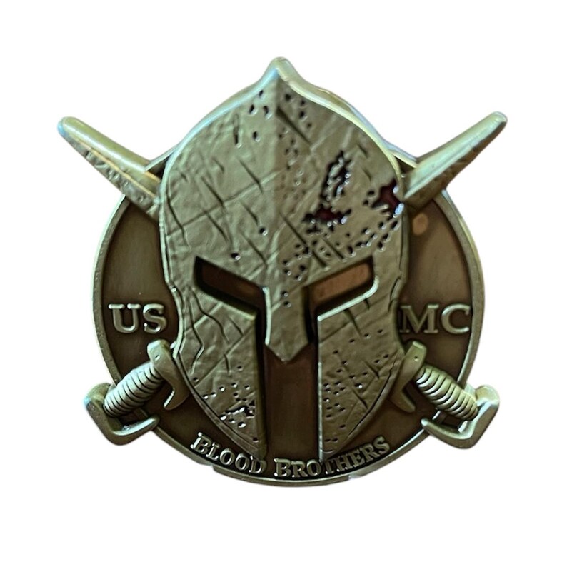 K&ouml;nnte beinhalten: Eine bronzefarbene M&uuml;nze mit einem Spartanerhelm-Design. Die M&uuml;nze tr&auml;gt die Aufschrift "US MC" und "BLOOD BROTHERS". Zwei gekreuzte Schwerter sind ebenfalls vorhanden. Die M&uuml;nze hat ein abgenutztes, gealtertes Aussehen, mit einem Durchmesser von ca. 4 cm.