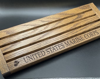 Puede incluir: Un expositor rectangular de madera con cuatro ranuras horizontales. Las palabras "UNITED STATES MARINE CORPS" están grabadas en la parte delantera, junto con el emblema de los Marines. La madera tiene un tono marrón cálido natural.