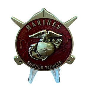 K&ouml;nnte beinhalten: Ein goldfarbenes Marine Corps-Emblem mit rotem Hintergrund. Das Emblem zeigt die Worte "MARINES" und "SEMPER FIDELIS" sowie einen Adler, einen Globus und einen Anker. Das Emblem wird auf einem transparenten St&auml;nder pr&auml;sentiert.